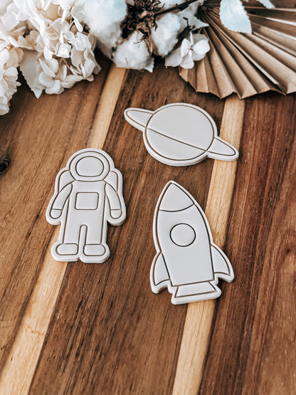 Wooden cutouts of an astronaut, planet, and rocket on a wooden surface with cotton and paper decorations.